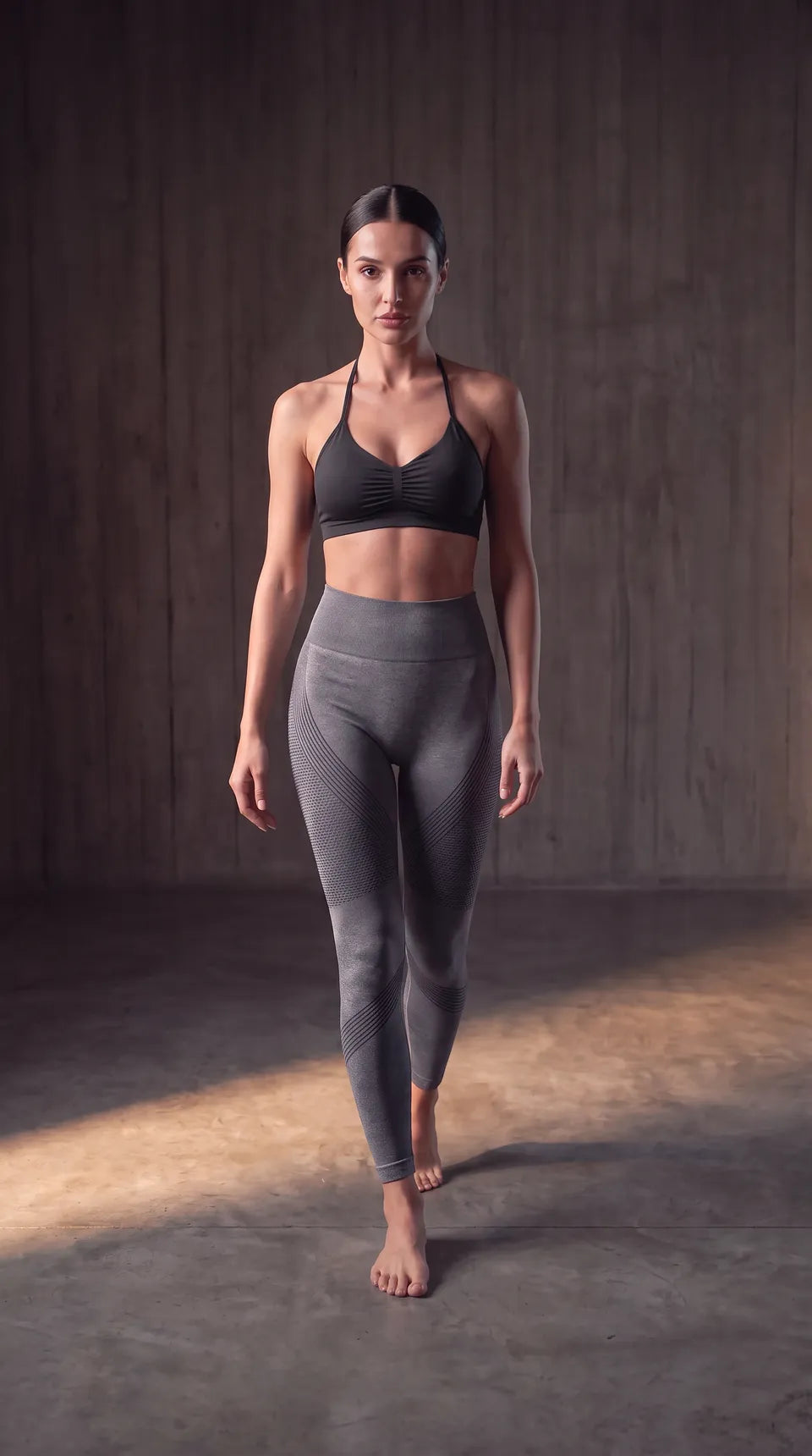Elle marche vers la caméra dans la bande de lumière — La Brassière Nouée noir et Le Legging Sculptant gris ardoise.